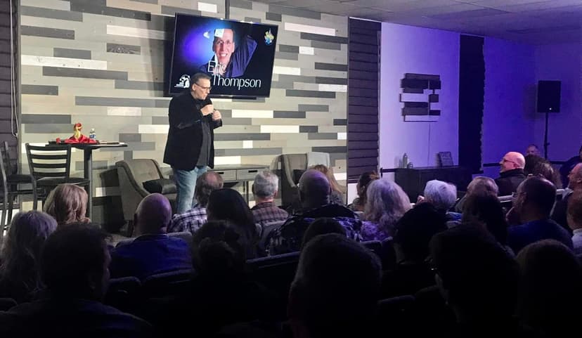 Eric Thompson commands the stage with his signature warmth and humor, captivating a packed audience in this intimate venue setting. The veteran entertainer works his magic on the engaged crowd, with his name proudly displayed on the screen behind him.