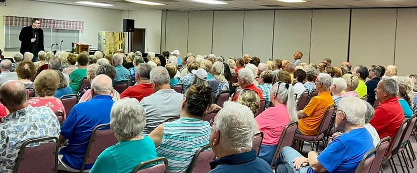 Eric Thompson captivates a packed audience during what appears to be a community or church event, with dozens of attentive faces hanging on his every word from the stage.