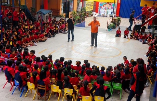 Eric Thompson captivates a sea of young students in matching red shirts during what appears to be an energetic school assembly performance, with the kids forming a perfect circle around the entertainer.