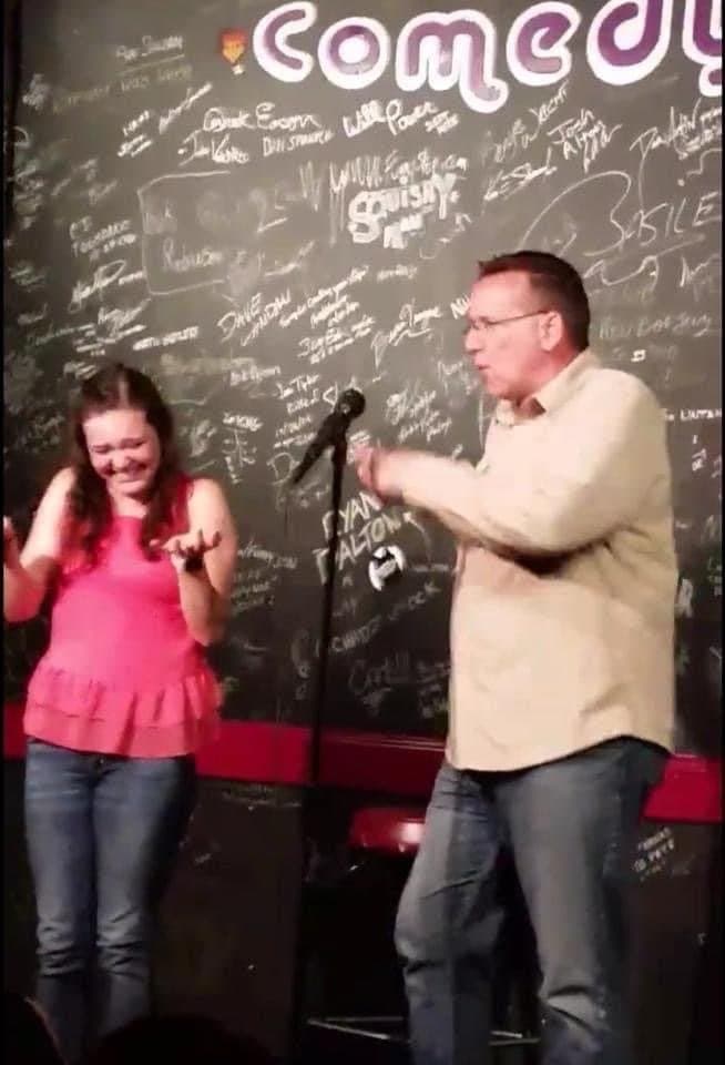 Eric Thompson brings a delighted audience member on stage for what looks like an interactive comedy bit, with her covering her face in laughter while he works his comedic magic at this intimate comedy club venue.