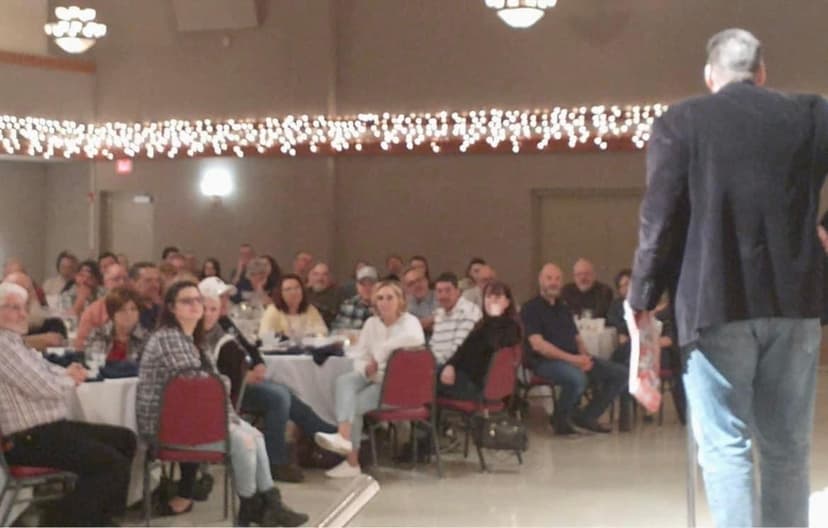 Eric Thompson commands the room at what appears to be a lively banquet hall event, with a captivated audience hanging on his every word as festive lights twinkle overhead.