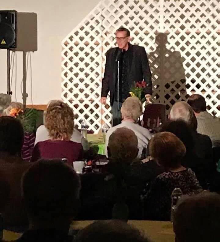 Eric Thompson commands the stage at an intimate venue, delivering his signature blend of comedy and magic to a captivated seated audience gathered around white lattice backdrop.