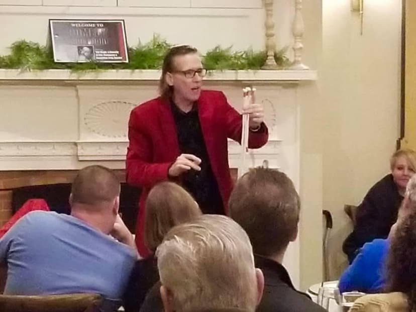 Eric Thompson captivates an intimate audience during what appears to be a private event or small venue performance, demonstrating his signature blend of comedy and magic with animated gestures.