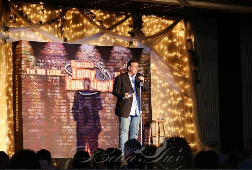 Eric Thompson takes the stage at an intimate comedy club, working his magic under the warm glow of twinkling string lights and that classic brick wall backdrop that every comedian knows and loves.