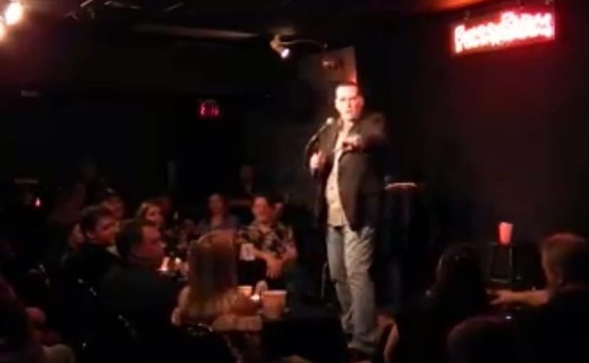 Eric Thompson commands the stage at an intimate comedy club, working his magic on a captivated audience packed around small tables in the dimly lit venue.