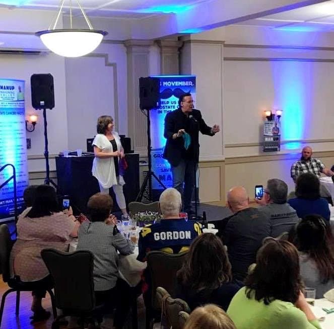 Eric Thompson commands the room at what appears to be a corporate event or fundraiser, engaging his seated audience with his signature blend of comedy and charm while a colleague looks on from the stage.