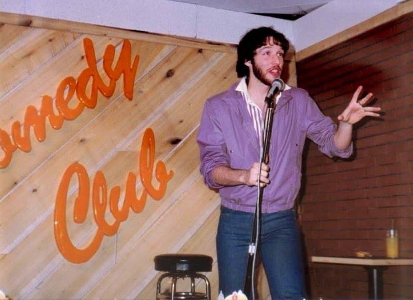 Eric Thompson commands the stage at what appears to be a cozy comedy club, gesturing expressively mid-performance as he delivers his signature blend of humor and entertainment to the audience.