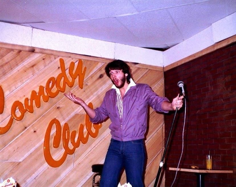 Eric Thompson commands the stage at what appears to be a comedy club, gesturing enthusiastically to the crowd with his signature animated style that has made him a favorite at venues across the country.