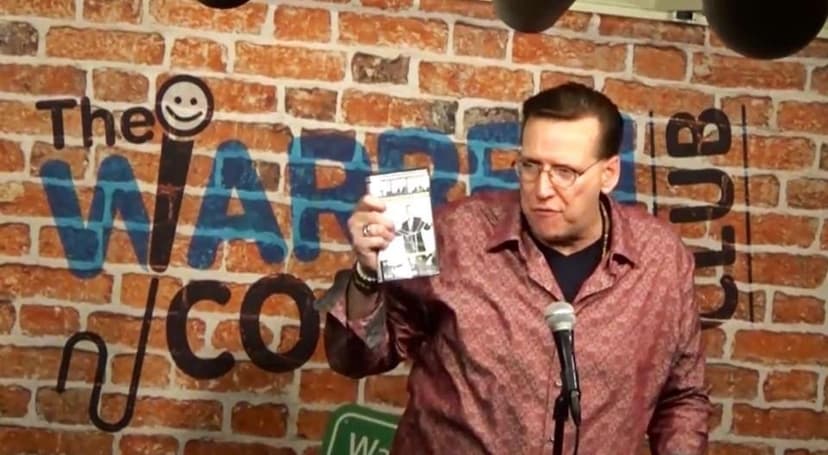 Eric Thompson holds up what appears to be a book or magazine during his engaging performance at a comedy club, sharing another hilarious moment with the audience against the iconic brick wall backdrop.