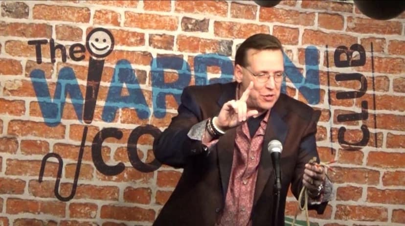 Eric Thompson brings his signature blend of comedy and charm to the stage at a comedy club, gesturing expressively while delivering what's clearly a crowd-pleasing bit in front of the classic brick wall backdrop.