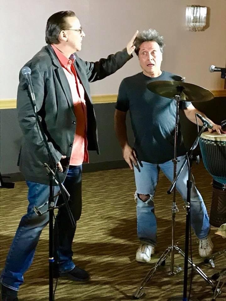 Eric Thompson shares a playful moment with a fellow performer, showing off his natural comedic timing as he playfully musses his colleague's hair during what appears to be a casual rehearsal or sound check.