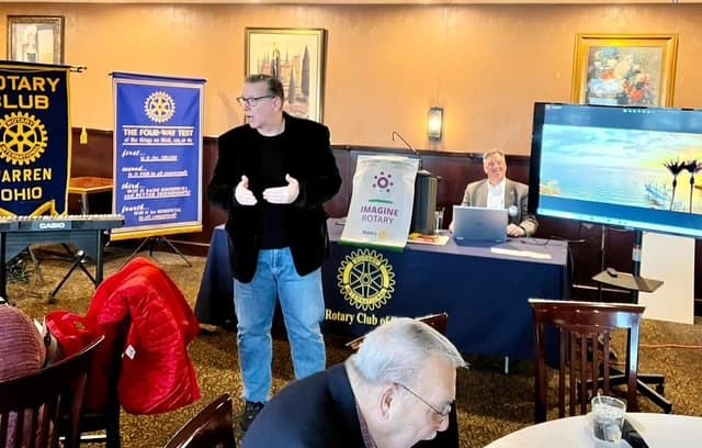 Eric Thompson brings his signature blend of comedy and magic to a Rotary Club meeting, captivating the audience with his animated storytelling while a fellow member looks on from behind the presentation table.