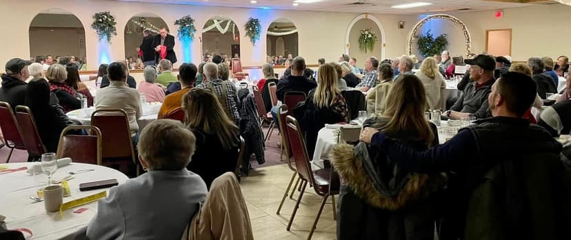Eric Thompson commands the stage at a packed banquet hall event, entertaining a captivated audience of dozens who are clearly enjoying every moment of his performance.