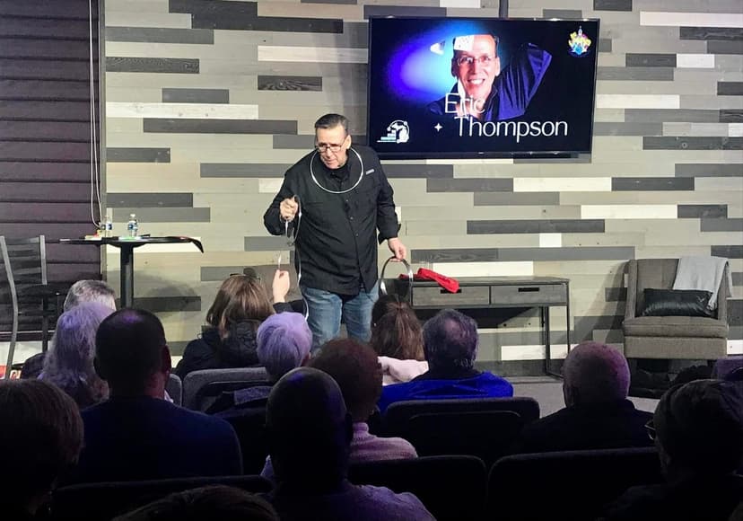 Eric Thompson captivates an intimate audience during what appears to be a corporate event or private show, working his magic in a modern venue with sleek wooden paneling.