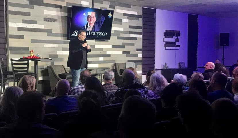 Eric Thompson commands the stage with microphone in hand, captivating a packed audience in this intimate venue setting. The seasoned entertainer works his magic on the attentive crowd while his image beams from the screen behind him.
