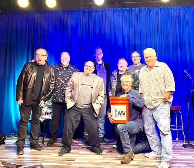Eric Thompson takes center stage with his comedy troupe for a group photo after what looks like a hilarious collaborative performance, complete with props and big smiles all around.