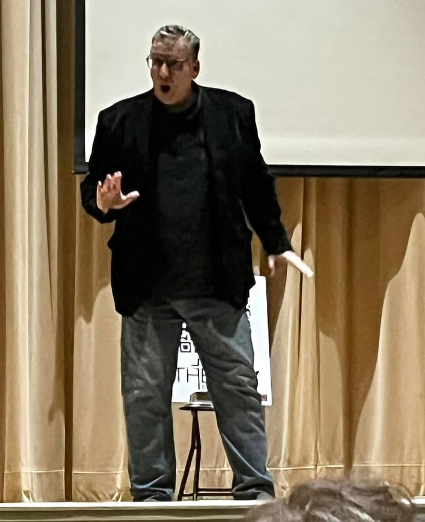 Eric Thompson commands the stage with animated gestures and expressive storytelling, captivating his audience with his signature blend of comedy and showmanship at what appears to be a speaking engagement.