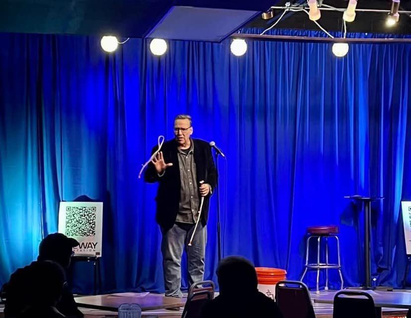 Eric Thompson commands the stage with his signature charm, gesturing expressively as he delivers his comedy routine under the bright stage lights at what appears to be an intimate comedy club performance.