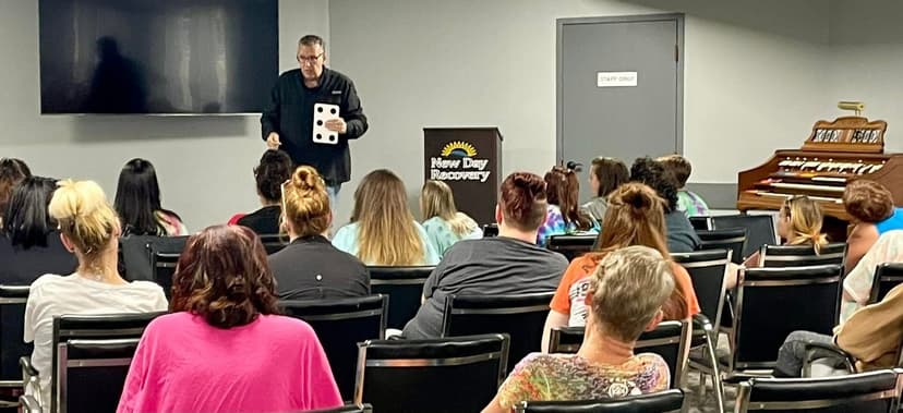 Eric Thompson captivates a packed audience at what appears to be a recovery center event, holding up oversized dice as part of his engaging performance routine.