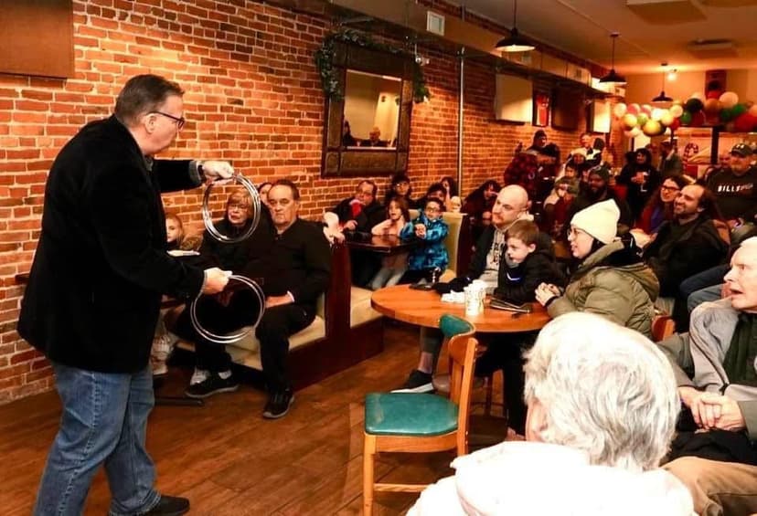 Eric Thompson captivates a packed audience at what appears to be a cozy restaurant or café venue, demonstrating his signature blend of magic and comedy with metal rings while the crowd watches in anticipation.