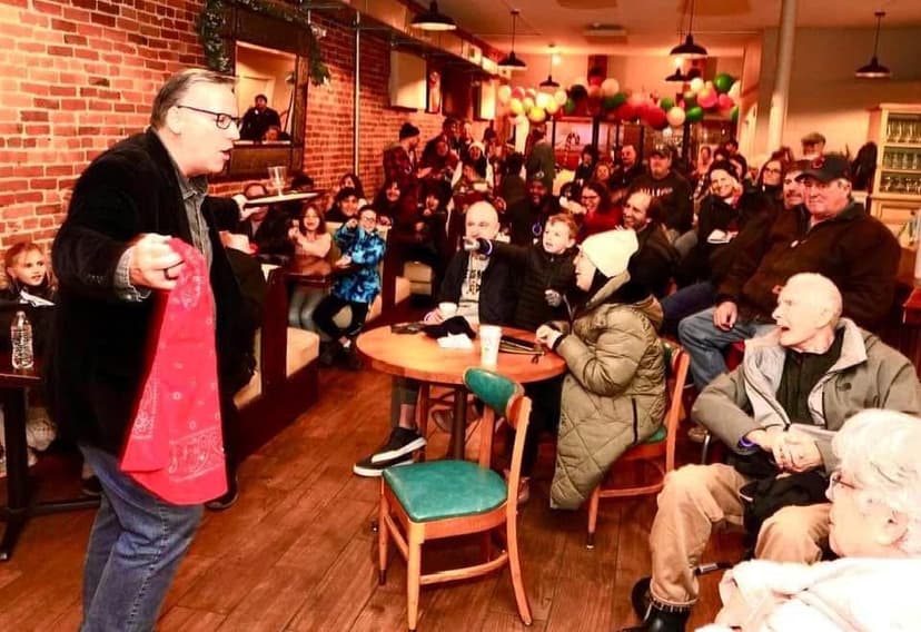 Eric Thompson captivates a packed house at what appears to be a holiday party or fundraiser, holding up a festive red item while his engaged audience watches intently from their seats.