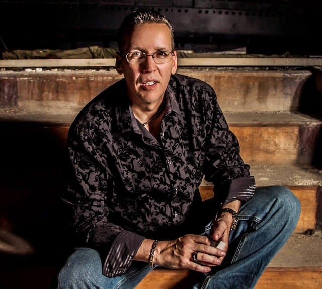 Eric Thompson strikes a relaxed pose during what appears to be a casual portrait session, showcasing his approachable showman style in a patterned shirt and jeans against a rustic stone backdrop.