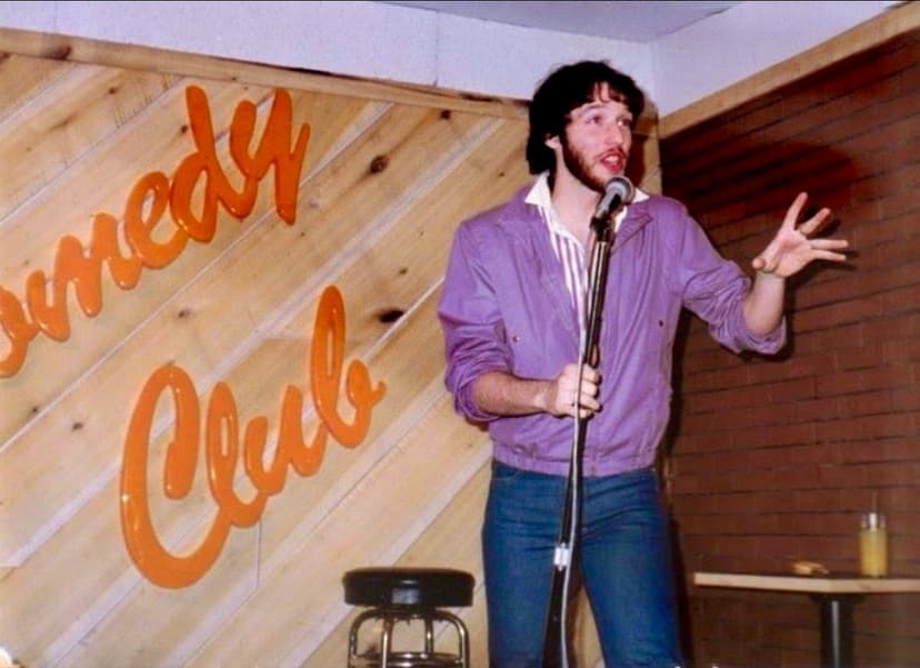 Eric Thompson takes the stage at what appears to be an intimate comedy club venue, gesturing expressively as he delivers his routine to the audience.