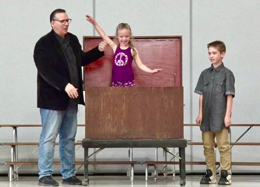 Eric Thompson delights a young audience member during his magic show, with the girl appearing to emerge from or disappear into a classic magician's box while another child watches the amazing trick unfold.