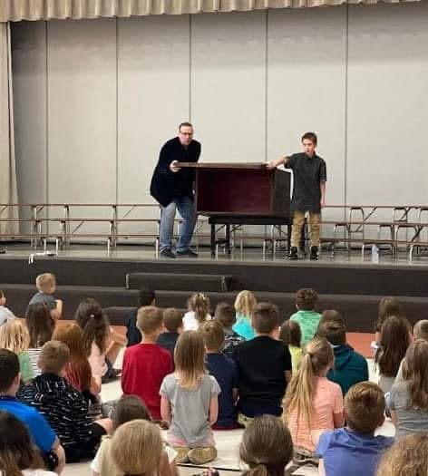 Eric Thompson captivates a young audience from the stage, working his magic and comedy charm on what appears to be a school assembly full of eager kids.