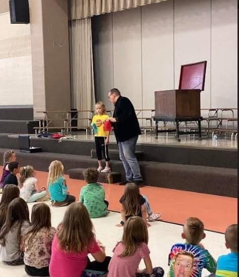 Eric Thompson captivates a group of young audience members during what appears to be a school or community center performance, with the kids sitting cross-legged and completely engaged as he works his magic on stage.