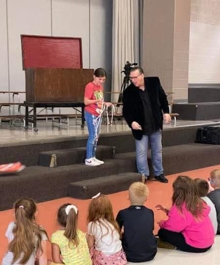Eric Thompson captivates a young audience during what appears to be a school or church performance, with children sitting cross-legged and completely engaged as he works his comedic magic.