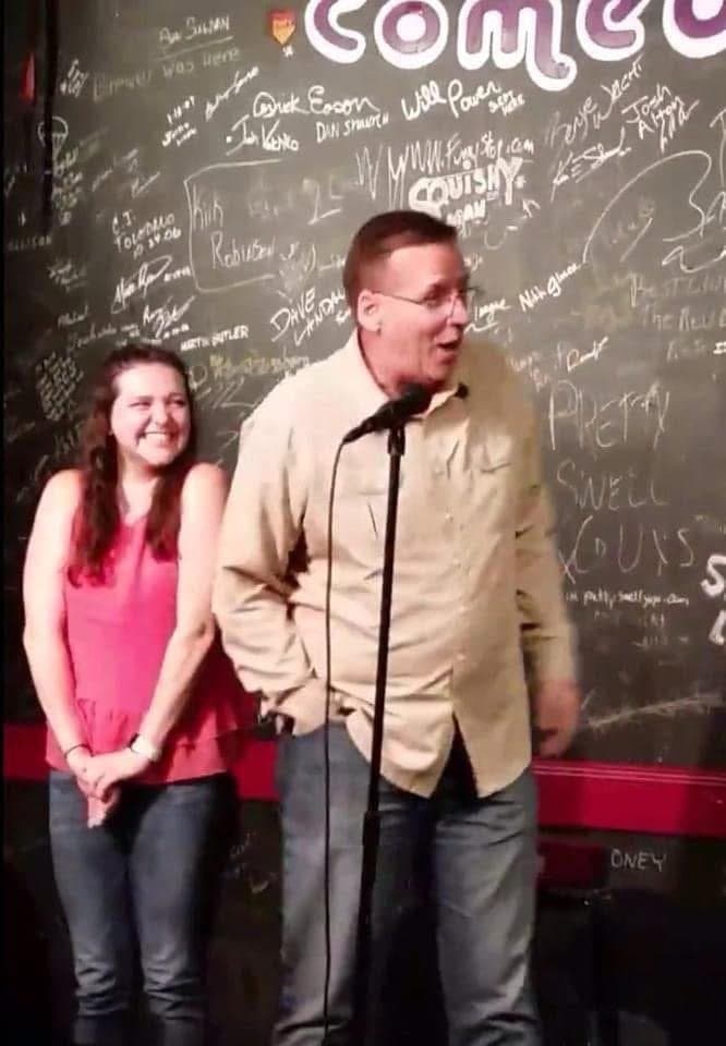 Eric Thompson shares the stage with an audience member at what appears to be a comedy club, both grinning as he works his comedic magic in front of a wall covered with performer signatures.