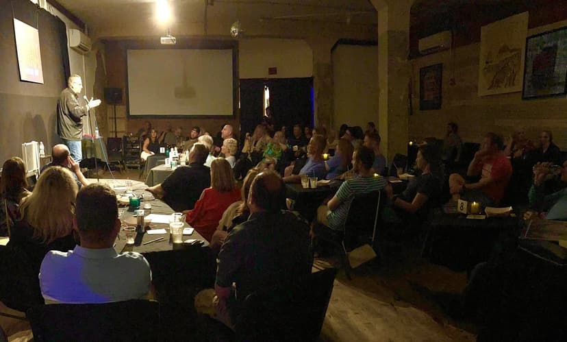 Eric Thompson commands the room during what appears to be a corporate event or fundraiser, with his audience seated at tables and clearly engaged in his entertaining presentation.