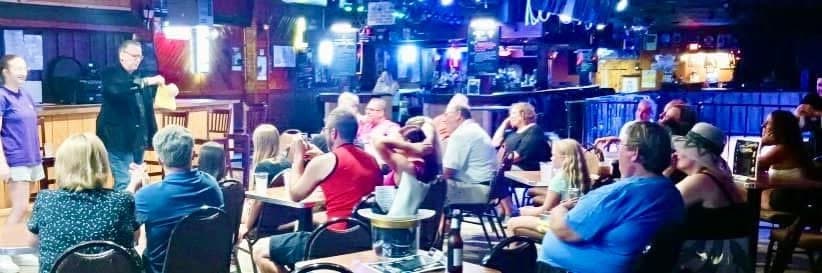 Eric Thompson captivates a packed audience at what appears to be a lively restaurant or comedy club venue, with dozens of guests seated at tables enjoying his entertaining performance under the warm stage lighting.