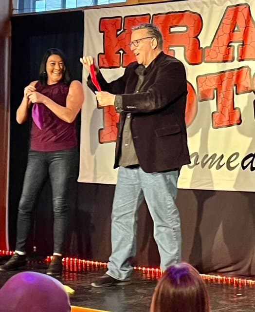 Eric Thompson brings a volunteer on stage for what looks like a hilarious magic trick or comedy bit, with both performer and participant sharing genuine laughter at this lively venue.