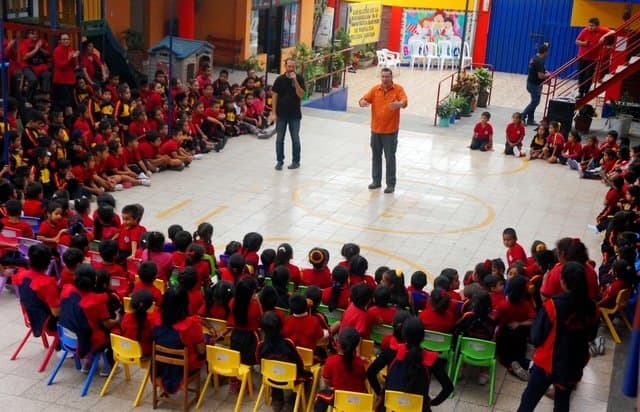 Eric Thompson captivates a packed audience of students in their bright red uniforms, commanding the center of the room as he delivers what's sure to be an unforgettable performance at this school assembly.