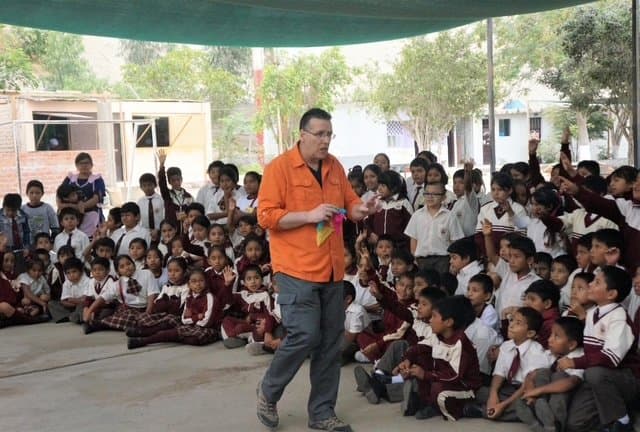 Eric Thompson captivates a sea of school children during what appears to be an educational assembly, his bright orange shirt making him impossible to miss as he works his magic with the enthusiastic young audience.