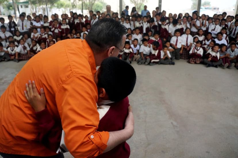 Eric Thompson shares a heartwarming moment with a young student during what appears to be a school assembly, with hundreds of uniformed children sitting cross-legged and watching the touching interaction unfold.