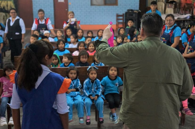 Eric Thompson captivates a room full of young students during what appears to be a school assembly, holding up a colorful prop while the kids watch in rapt attention from their seats.
