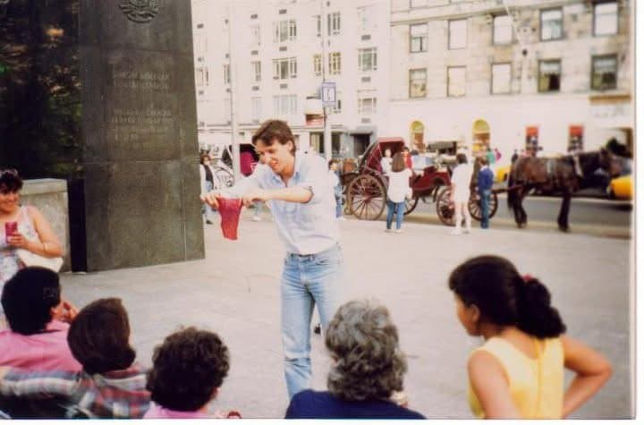 Eric Thompson captivates a seated audience with his street performance magic in what appears to be a charming European town square, complete with horse-drawn carriages adding to the old-world atmosphere.