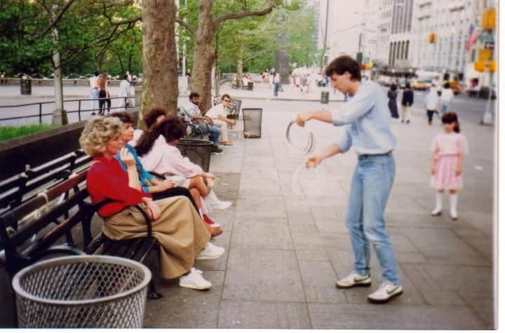 Eric Thompson captivates an outdoor audience with his street performance magic, drawing curious onlookers as he works his comedic charm in what appears to be a bustling city plaza.
