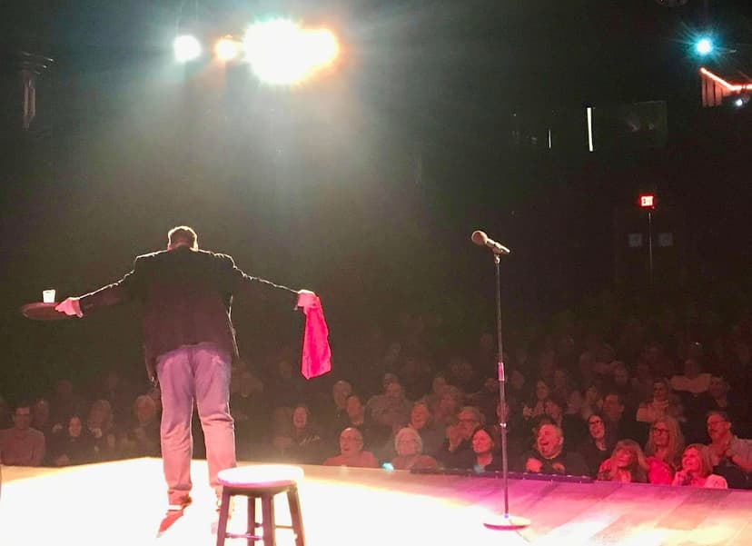 Eric Thompson commands the stage with theatrical flair, arms outstretched as he works his magic before a captivated theater audience bathed in dramatic stage lighting.