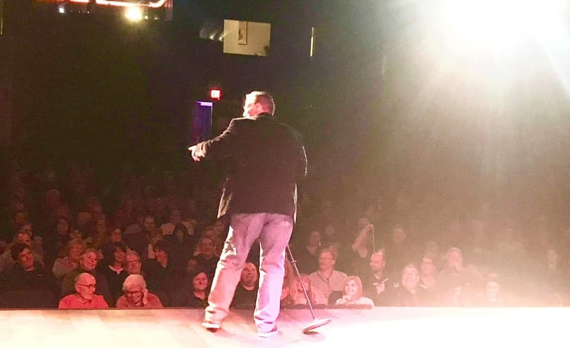 Eric Thompson commands the stage in dramatic lighting as he captivates a packed theater audience, his animated gestures and energy filling the venue with anticipation.