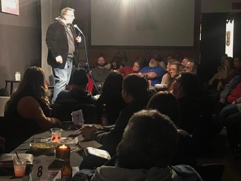 Eric Thompson commands the stage at an intimate comedy club, delivering his signature blend of humor to a packed house of appreciative fans seated at cozy dinner tables.