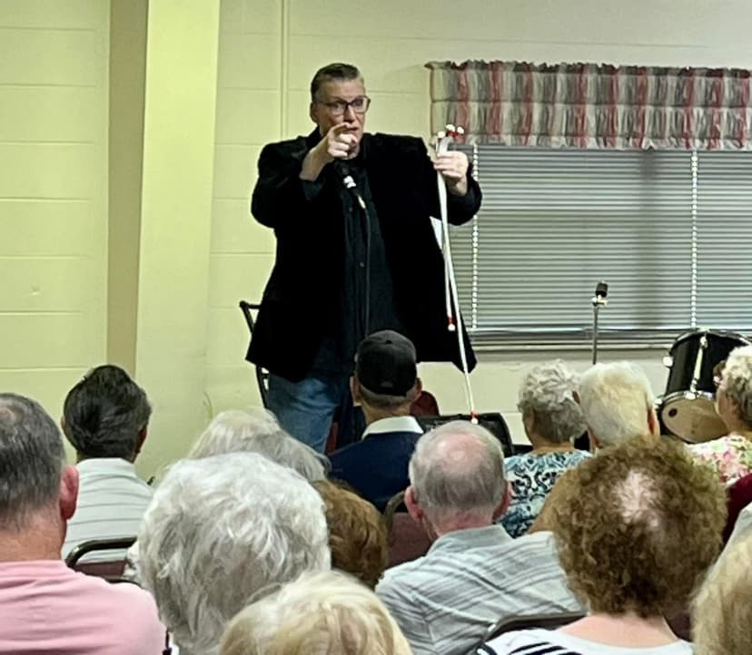 Eric Thompson captivates a packed audience during what appears to be a community center or church performance, gesturing animatedly as he delivers his signature blend of comedy and magic to an engaged crowd of all ages.