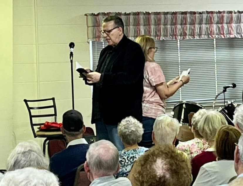 Eric Thompson captivates an intimate audience during what appears to be a church or community center performance, reading from his material while a blonde assistant stands ready nearby.