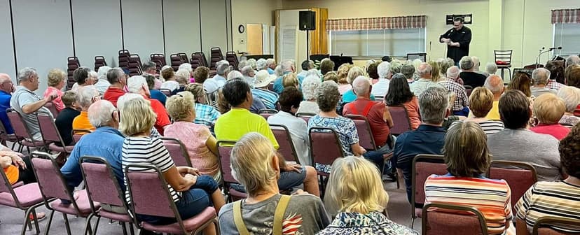 Eric Thompson commands the attention of a packed audience in what appears to be a community center or church hall, demonstrating his ability to draw and engage crowds of all ages with his signature blend of comedy and magic.
