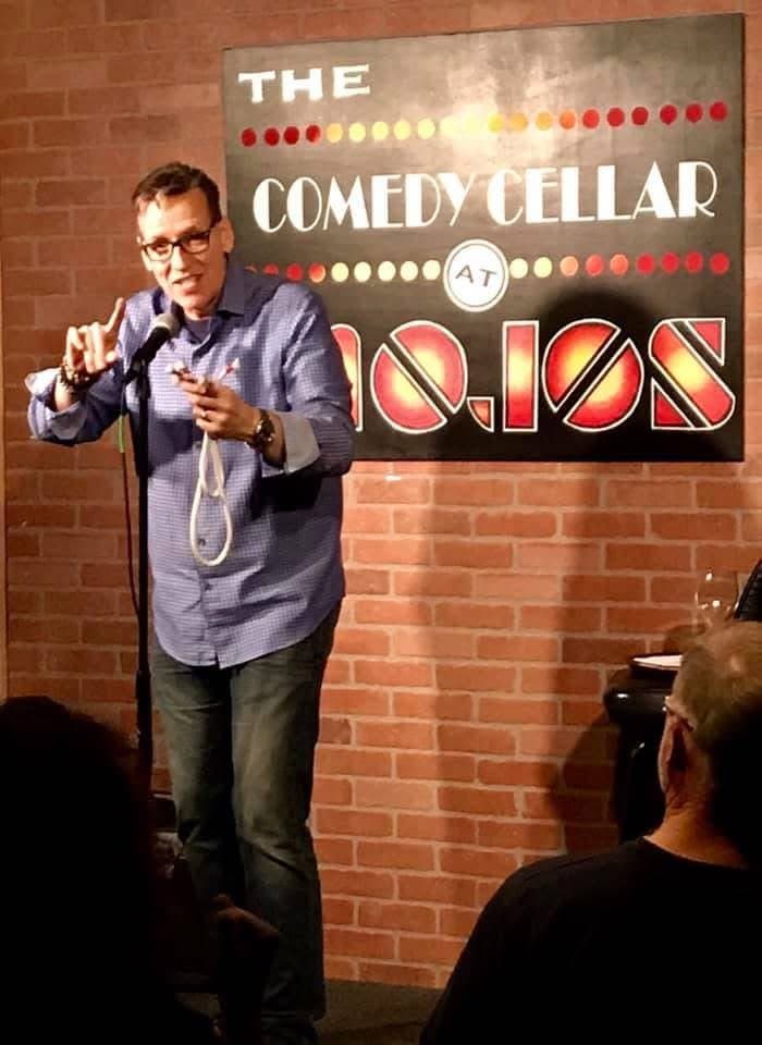 Eric Thompson commands the stage at The Comedy Cellar, gesturing confidently as he delivers his signature blend of comedy and magic to an intimate crowd.