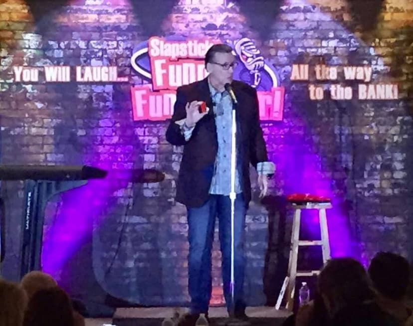Eric Thompson takes the stage at a comedy club, working his magic with the microphone as purple stage lights create the perfect atmosphere for an evening of laughs. The veteran entertainer commands the brick-walled stage with his signature blend of comedy and showmanship.
