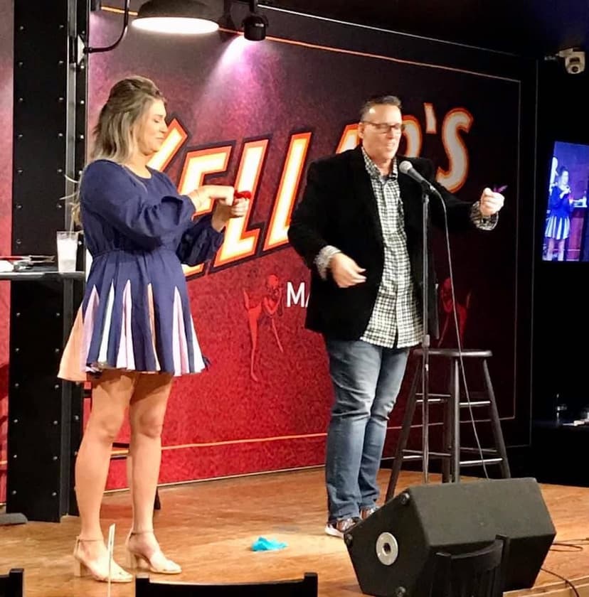 Eric Thompson charms the audience at what appears to be a comedy club performance, sharing the stage with a woman in a striking blue dress as they work together during his entertaining set.
